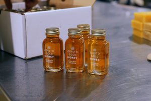Four 3-ounce glass bottles of Edisto Gold Honey with gold caps arranged on a stainless steel counter next to a white box.