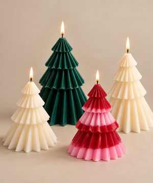 Three tiered Christmas tree-shaped candles on a warm beige background, including one dark green candle, one cream candle, and one ombre candle transitioning from red to pink.