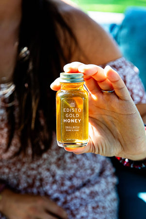 A hand holding a 3-ounce glass bottle of Edisto Gold Honey with a gold cap, shown against a blurred background of a person wearing a patterned top.