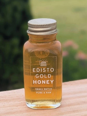 A 3-ounce glass jar of Edisto Gold Honey with a gold metal cap, sitting on a wooden surface with a blurred green outdoor background.