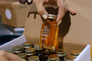 A hand holds a 3-ounce glass bottle of Edisto Gold Honey, positioned above an open white box containing multiple similar honey bottles.